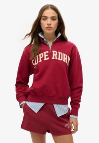 Rode zip-up sweatshirt met "SUPERDRY" logo, geribde manchetten en zoom; gecombineerd met een blauwe gestreepte shirt die eronderuit steekt en bijpassende shorts.