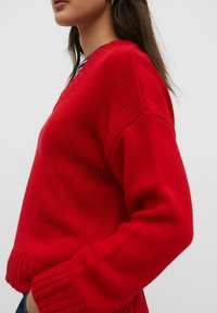 Rode gebreide trui met een V-hals, geribde manchetten en onderrand. Beschikt over een gestructureerd patroon en een relaxte pasvorm.