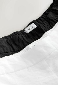 Două perechi de pantaloni scurți, una neagră și una albă, cu talie elastică. Etichetele indică "NEXT." Material din bumbac cu textură netedă.