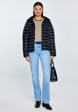 Nő fekete puffert visel, csíkos inget, világoskék farmert, fekete csizmát, fekete kézitáskát tart, sima háttér előtt áll.