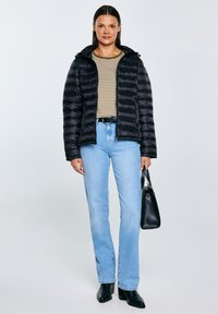 Vrouw draagt een zwarte donzen jas, gestreept shirt, lichtblauwe spijkerbroek, zwarte laarzen, houdt een zwarte handtas vast, staat voor een effen achtergrond.