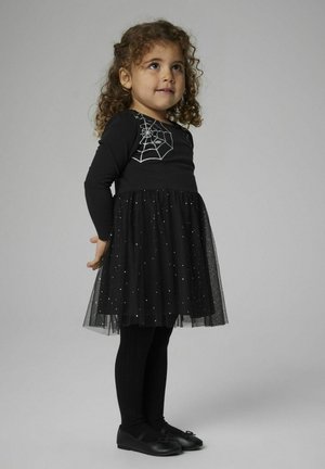 Schwarzes Kleid mit langen Ärmeln, das ein silbernes Spinnennetzdesign zeigt, Tüllrock mit silbernen Sternelementen, kombiniert mit schwarzen Strumpfhosen und Schuhen.