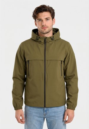 Uomo che indossa una giacca con cappuccio verde oliva con cerniera frontale e tasche sul petto con zip, abbinata a jeans azzurri, in piedi su sfondo bianco.