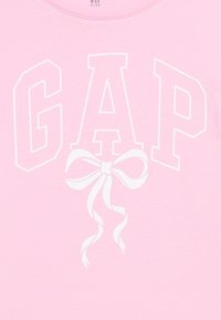 Ροζ ύφασμα με λευκά γράμματα "GAP" με περίγραμμα και ένα λευκό γραφικό φιόγκο στο κέντρο κάτω από το "A", μέρος ενός παιδικο ύ ρούχου.