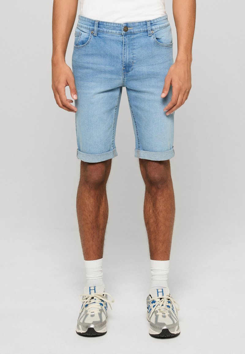 Mężczyzna ubrany w jasnoniebieskie, podwinięte krótkie spodenki dżinsowe, białe skarpetki i białe buty sportowe z niebieskimi elementami, stojący na jednolitym tle.