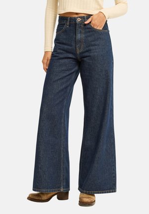Femme portant un jean bleu foncé taille haute à jambes larges avec coutures visibles, main dans la poche, pull court crème et bottines marron à talons.