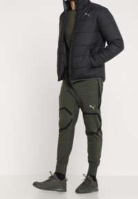 Man med svart vadderad jacka, olivgröna Puma joggers med svarta detaljer och matchande mörkgröna sneakers, står med händerna i fickorna.