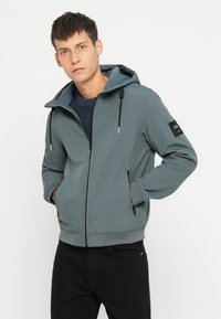 Hoodie in Schiefergrau mit einem Frontreißverschluss. Verfügt über verstellbare Kordelzüge, zwei Seitentaschen und ein schwarzes Akzentpatch am Ärmel.