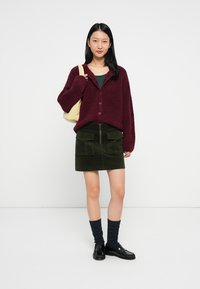 Cardigã peludo bordeaux com botões, blusa verde justa, saia mini de veludo cotelê azeitona com bolsos, meias até o joelho azuis e mocassins pretos.