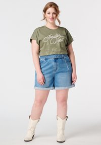 Olijfgroen t-shirt met "Jardin Vert" opdruk, gecombineerd met lichtblauwe denim shorts met opgerolde zoom. Witte cowboylaarzen maken de look af.