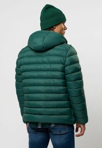 Groene pufferjack met capuchon, geborduurde textuur en een gladde afwerking. Draagt over een geruite blouse met een relaxte pasvorm en blauwe jeans.