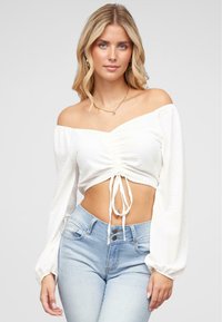 Bílý crop top s odhalenými rameny, se sklady vpředu a detailem na zavázání, spojený s světle modrými džíny s vysokým pasem. Hladká tkanina, balonové rukávy.