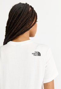 Tricou alb cu mânecă scurtă, având un logo negru "The North Face" pe partea superioară a spatelui. Materialul pare neted și ușor.