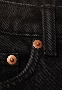 Κοντινή λήψη μαύρου τζιν υφάσματος με χάλκινο κουμπί με χαραγμένο κείμενο, ενισχυμένη ραφή και επίπεδη μέση.