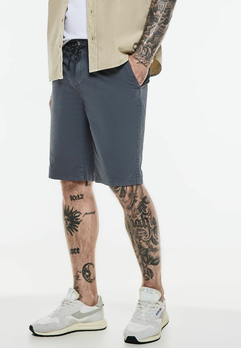 Shorts gris en tissu léger, dotés d'une taille élastique avec cordon de serrage, de poches latérales et d'une coupe droite, associés à des baskets blanches.