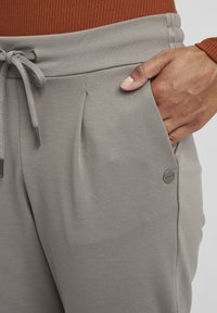 Grå joggebukser med snor i livet, fold og sidelommer. Har en liten sølvlogo detalj og myk, teksturert stoff.