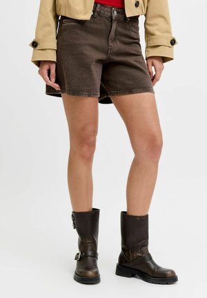 Femeie purtând pantaloni scurți din denim maro închis, jachetă bej și cizme din piele maro închis până la mijlocul gambei cu catarame, stând pe un fundal alb.