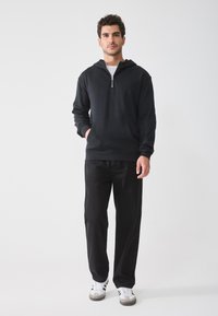 Sudadera negra con cremallera, bolsillo tipo canguro y mangas largas, combinada con pantalones negros y zapatillas blancas con detalles en negro. Un look simple y casual.