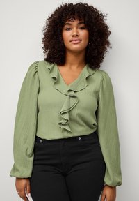 Blouse verte texturée avec un col froncé et des manches ballon longues, portée avec un pantalon noir taille haute.