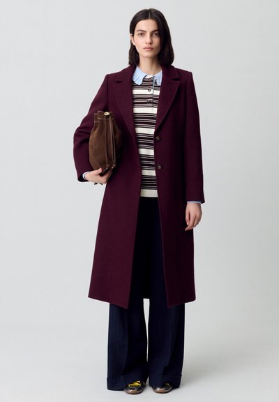 Abrigo de lana burdeos con cuello de muesca, combinado con un suéter a rayas y pantalones de pierna ancha en azul marino. Sosteniendo un bolso de ante marrón.