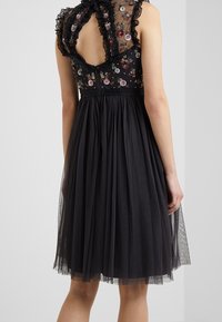 Robe noire avec un corsage en dentelle transparent brodé présentant des motifs floraux. La jupe est en tulle superposé, avec une taille ajustée et un col à volants.