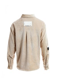 Camisa de pana beige con mangas largas, detalle de pliegues en la espalda y una etiqueta blanca en la parte superior de la espalda. Puños abotonados con un acabado texturizado.