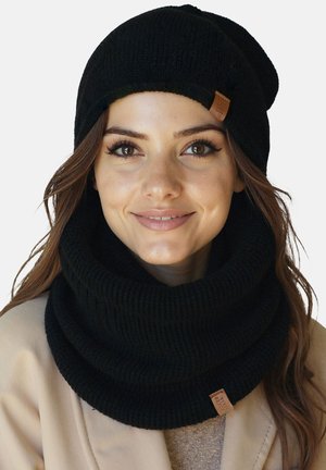 Žena se usmívá, má na sobě černou pletenou čepici a k ní ladící hustý černý pletený nákrčník přes béžový kabát.