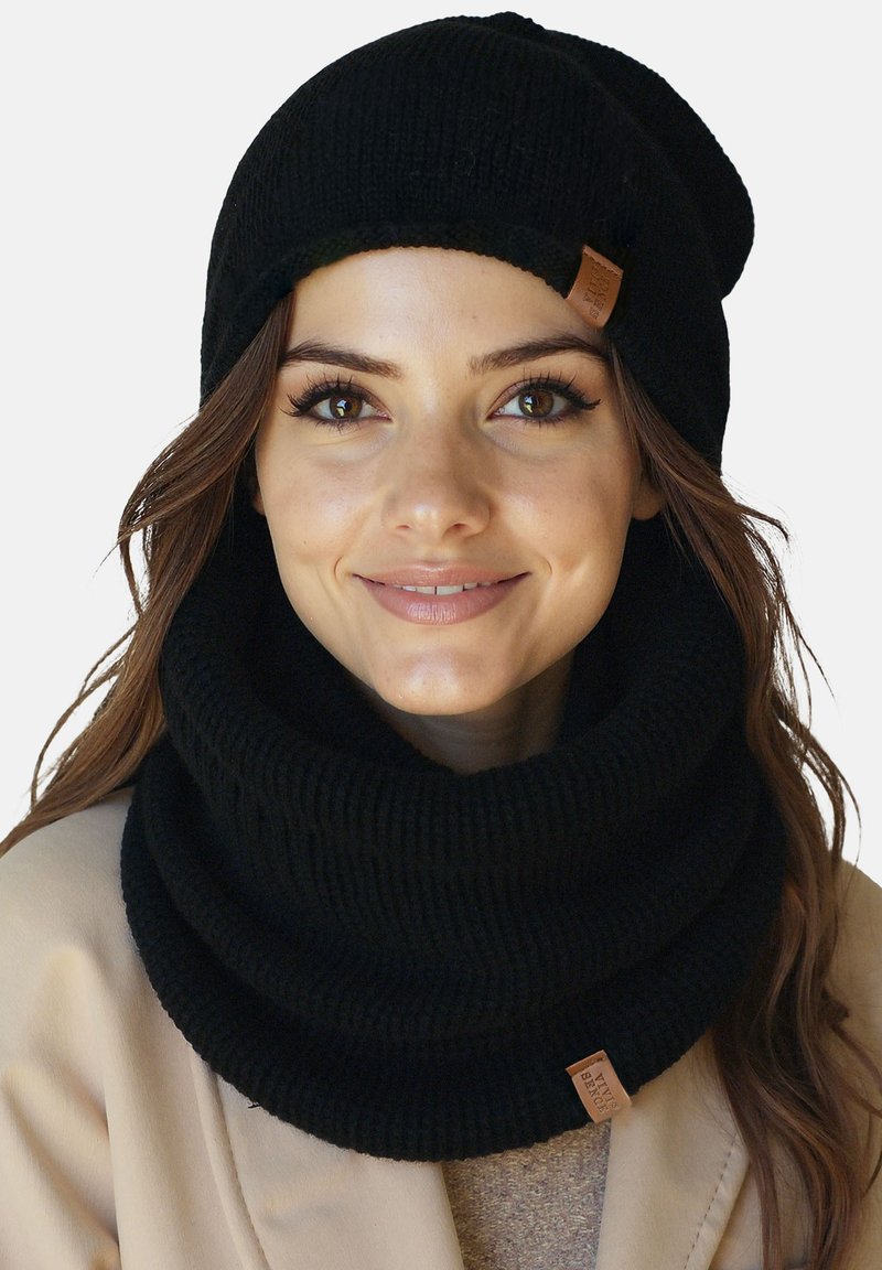 Mujer sonriendo, con un gorro de punto negro y un grueso calentador de cuello de punto negro a juego, sobre un abrigo beige.