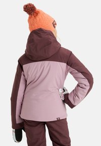 Paarse en bruine skijas met een capuchon, met lange mouwen en elastische manchetten; draagt een oranje gebreide beanie met een pompon.