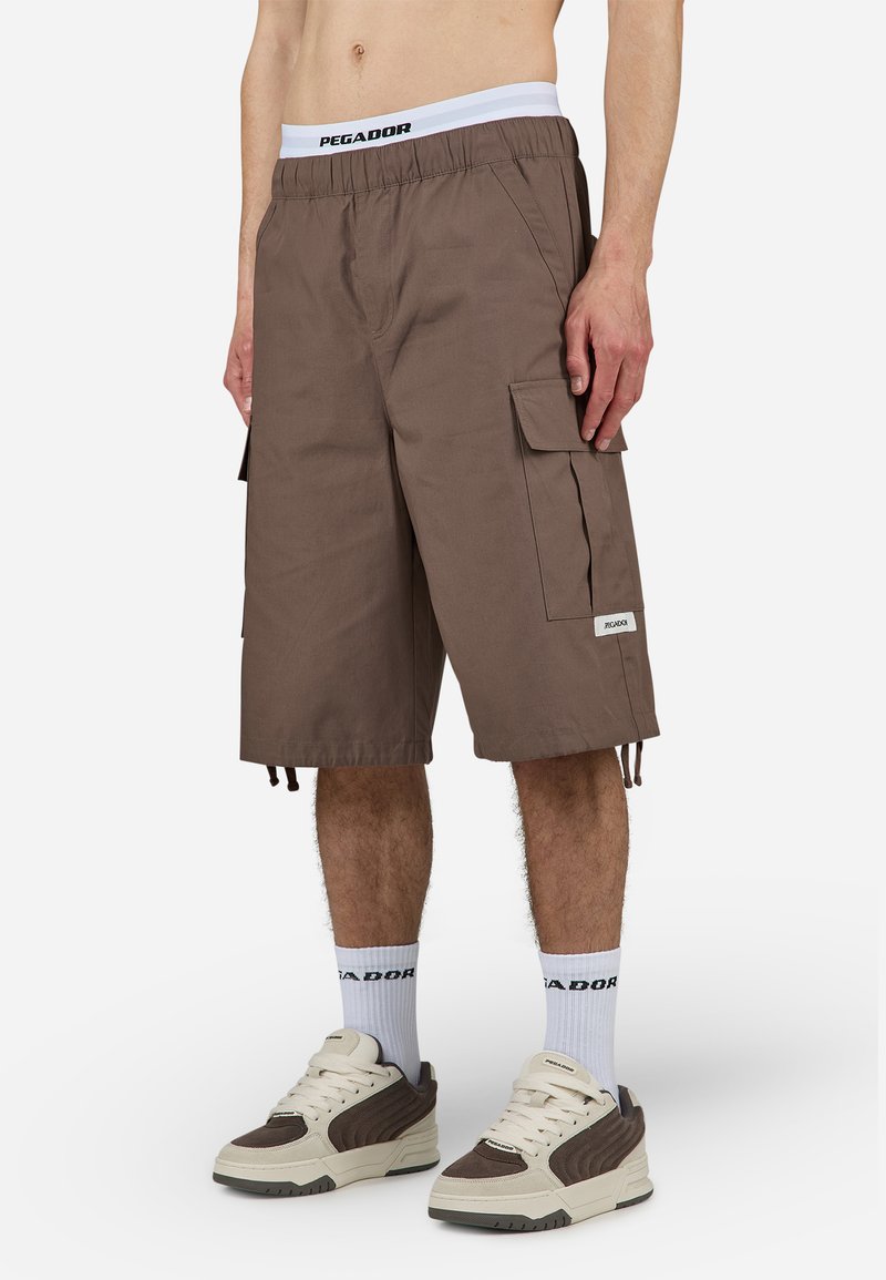 Brune cargo-short med elastisk waistband som har en hvit logo. Inkluderer flere sidelommer og justerbare snorer i kanten.