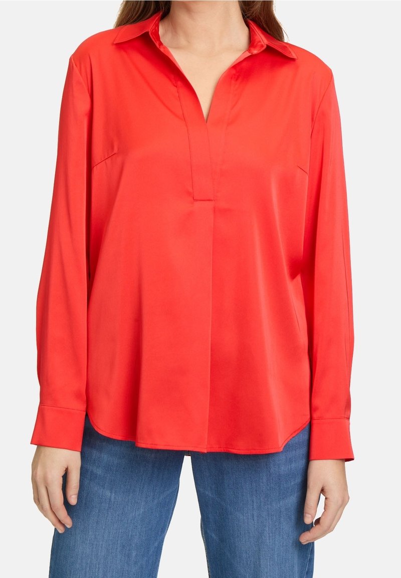 Blouse rouge vif à manches longues avec col et patte de boutonnage cachée, portée avec un jean bleu, montrée du cou jusqu'aux hanches.