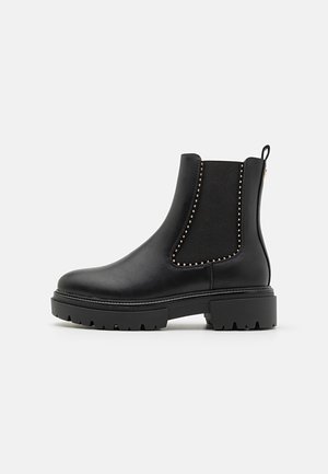 Botas Chelsea pretas com painéis laterais elásticos, sola de borracha texturizada, detalhes em tachas douradas e alça na parte de trás. Material de couro suave, altura até o tornozelo.