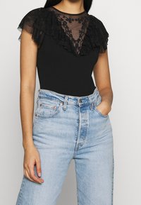 Czarny body z prześwitującym czarnym falbankowym dekoltem i kwiatową haftem, zestawiony z jasnoniebieskimi, wysokim stanem jeansami.