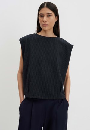 Femme aux longs cheveux foncés portant un haut noir sans manches avec de fines rayures verticales et un pantalon large sombre, les mains dans les poches.