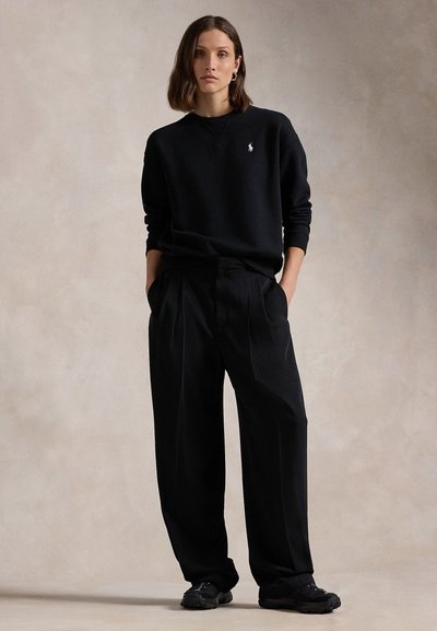 Küps naine, kelle pruunid juuksed ulatuvad õlgadeni, kannab musta logo jahedat sweatshirt'i, laia musta pükst ja musti spordijalatsid, seistes käed taskutes.