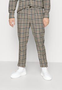 Rutigt plagg i brunt, svart och vitt mönster; långärmad skjorta och avsmalnande byxor, matchat med vita skor.