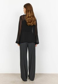 Black top with sheer long sleeves, paired with loose, dark grey trousers. Smooth fabric texture with a fitted bodice and relaxed fit.