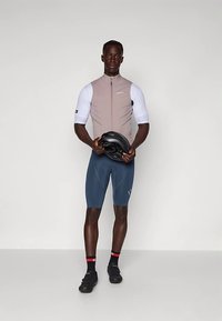 Mannlig syklist iført en beige ermeløs vest, hvit kortermet baselag, blå sykkelshorts, svarte sko, og holder en svart hjelm.