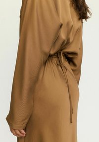 Hnědress s dlouhými, splývavými rukávy a staženým pasem se šňůrkou. Tkanina má jemný, texturovaný vzhled.