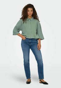 Camicia button-up verde chiaro, con texture, colletto e maniche ampie, abbinata a jeans blu slim-fit e ballerine nere.