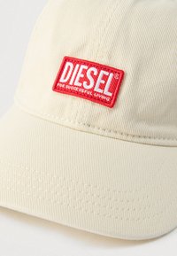Béžová bavlnená šiltovka s červenou obdĺžnikovou nášivkou s logom Diesel a nápisom "For Successful Living" na prednej časti.