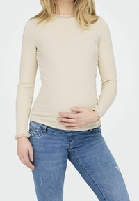 Beige långärmad topp med ribbad textur, med en volangkrage och manschetter, kombinerad med tighta blå denimjeans.