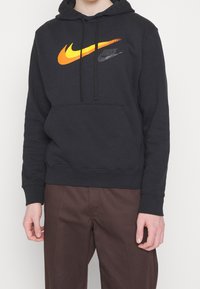 Svart hoodie med en känguruficka. Har en stor gradient Nike-logotyp i orange till gul och en mindre svart Nike-logotyp. Mjuk tygtextur.