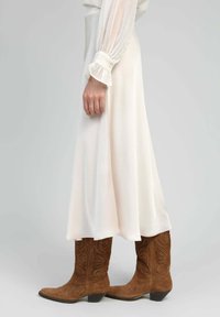 Robe en satin blanc avec de longues manches bouffantes et des poignets froncés, associée à des bottes de cowboy en daim marron ornées de motifs en relief.