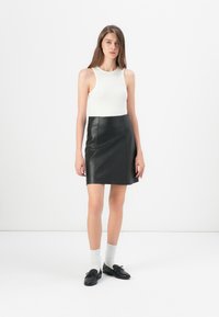 Schwarzer Lederrock mit leicht A-Linien-Form, kombiniert mit einem enganliegenden weißen Ripp-Top. Das Model trägt schwarze Schuhe und hellgraue Socken.
