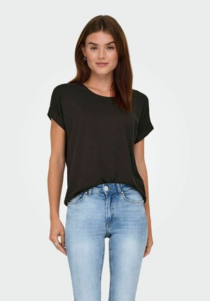 Woman with straight brown hair wearing black short-sleeve shirt and light blue jeans, standing against plain white background.