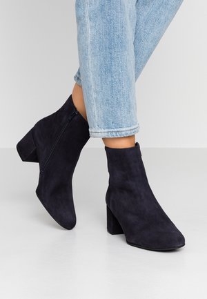 Tmavě modré semišové kotníkové boty s blokovým podpatkem, s postranním zipem a hladkou texturou. Nošeny s vyhrnutými světle modrými džínami.