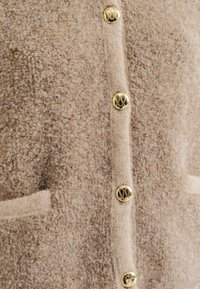 Beige, teksturert ullkåpe med en myk, fluffy overflate. Har gullknapper og to frontlommer, noe som skaper et klassisk design.