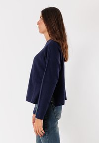 Donna con lunghi capelli castani che indossa un maglione a coste blu navy e jeans blu, in profilo contro uno sfondo bianco.