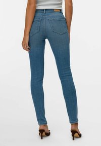 Peened sinine teksapaar, millel on keskmise kõrgusega vöökoht, viie-tasku disain ja tagaosa detailid. Kantud pruuni kõrge kontsaga kingadega.